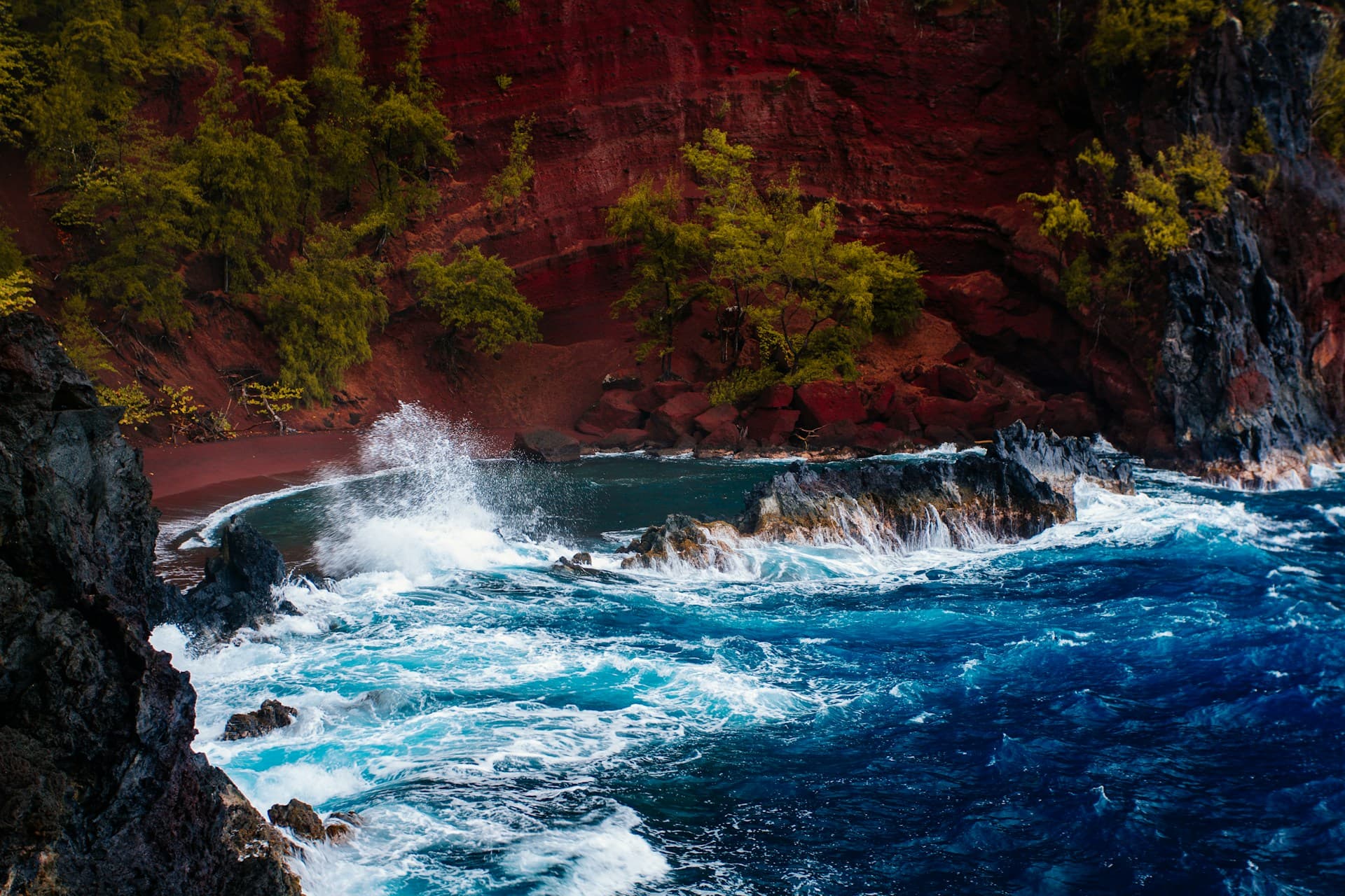 The deep red sand of Kaihalulu Beach in Hana Maui with turquoise water in the sheltered cove below volcanic cliffs